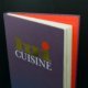 Un livre violet intitulé « Lui Cuisine » avec un point rouge sur la couverture, partiellement ouvert sur un fond noir, présente une superbe illustration et une touche de design graphique.