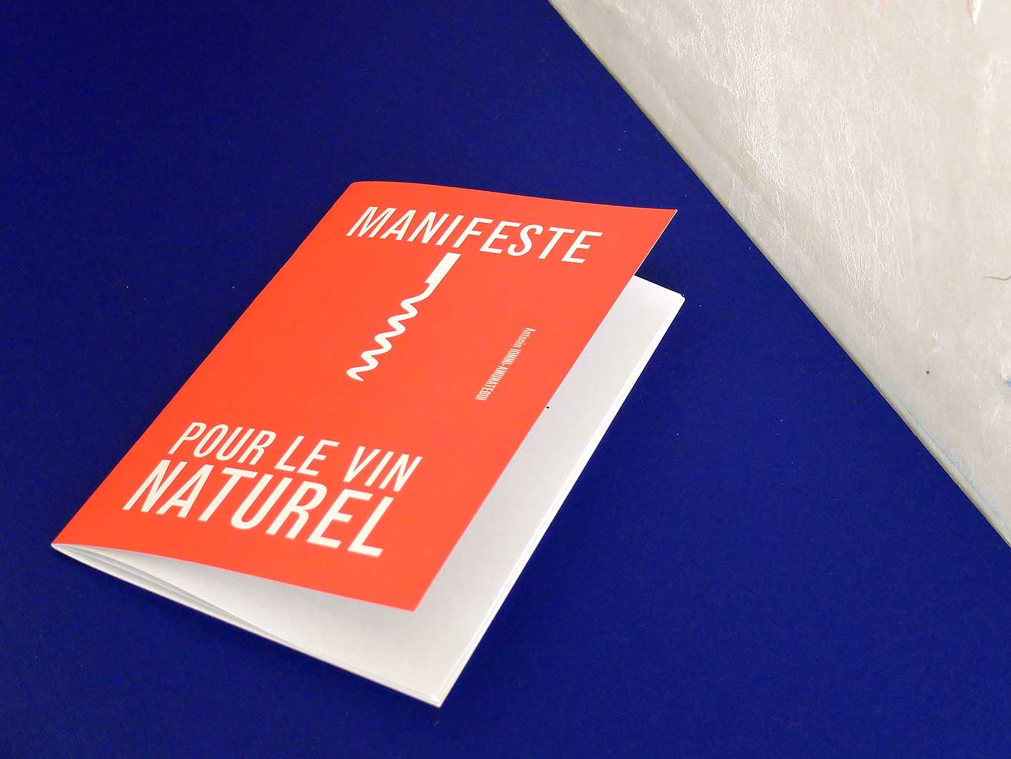 Un livret rouge intitulé « Manifeste pour le vin naturel » repose élégamment sur une surface bleue et blanche, se mariant harmonieusement avec son design graphique minimaliste.