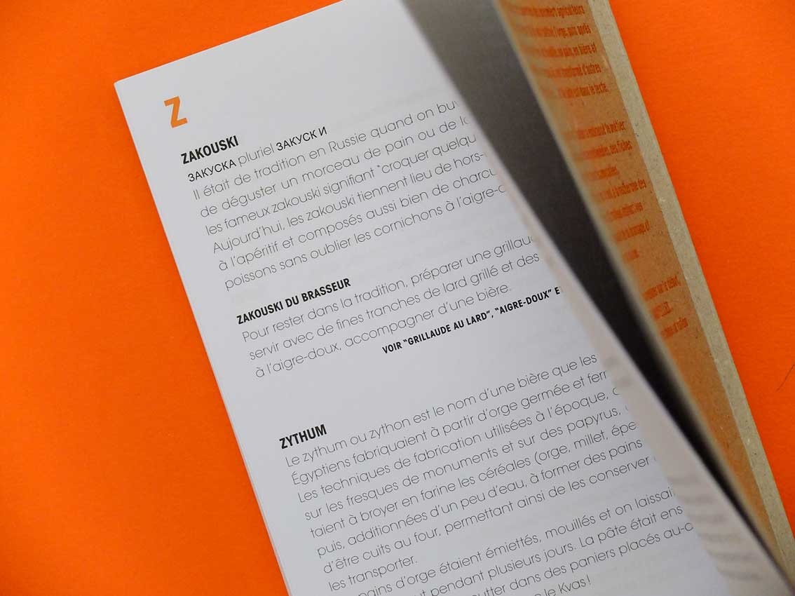 Un livre ouvert sur une surface orange présente des entrées de texte pour « Zakouski » et « Zythum », magnifiquement accompagnées de descriptions françaises illustratives. Les pages sont légèrement tournées, ajoutant une touche de design graphique à la scène.