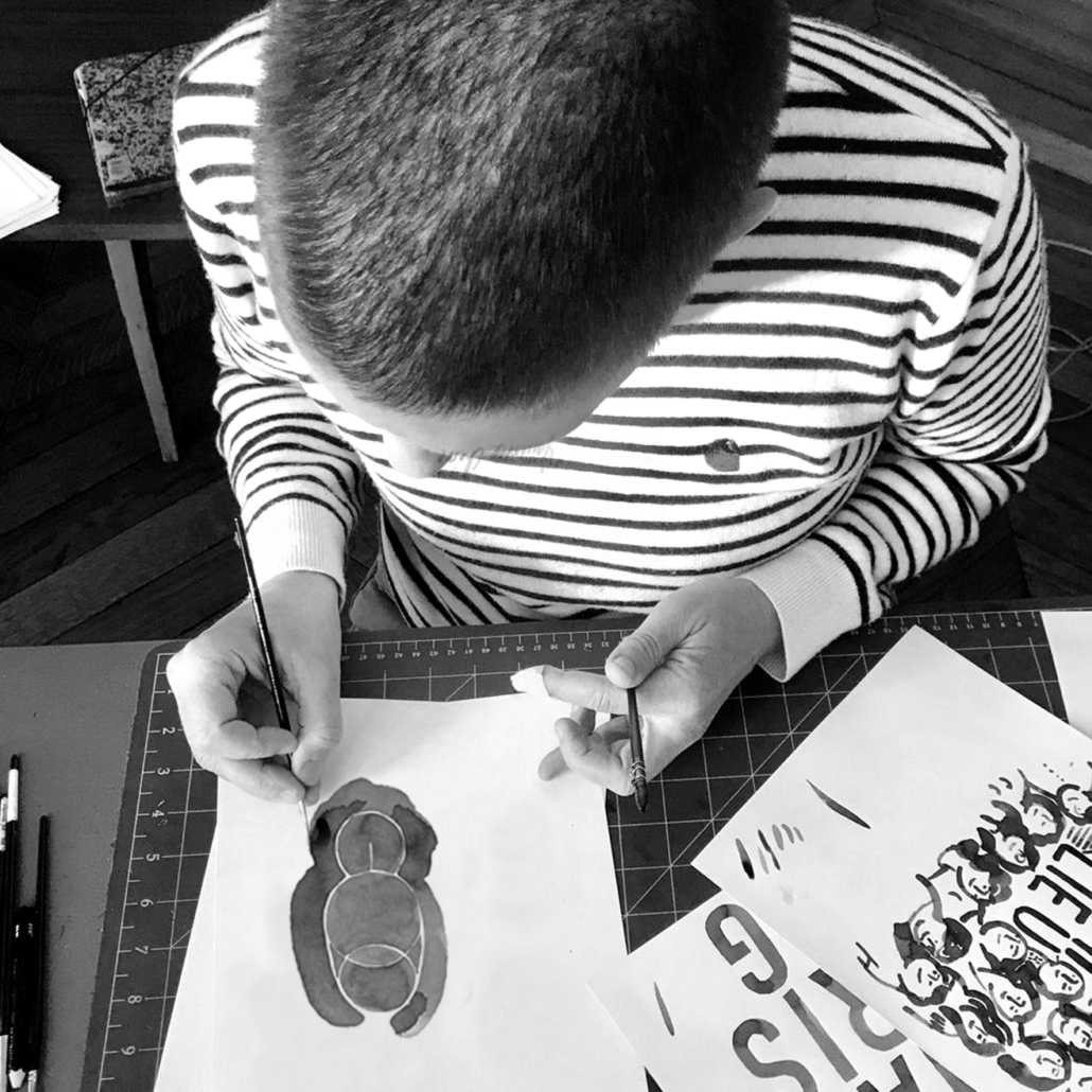 Laurence Chéné, en chemise rayée, peint sur du papier à un bureau orné de fournitures d'art, de croquis et d'un livre bien feuilleté à proximité.