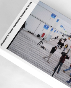 Un groupe de personnes munies d'appareils photo se tient près de bâtiments modulaires blancs portant des numéros bleus, que l'on aperçoit à travers une clôture métallique noire.